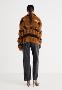 Femme avec une longue queue de cheval foncée portant un pull marron pelucheux rayé noir, un pantalon noir ample et des talons hauts imprimés léopard, debout de dos.