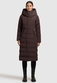 Brūns, garš pūkains mētelis ar augstu apkakli un lielu kapuci. Raksturīga horizontāla šūšana un rāvējslēdzēja sānu kabatas. Valkāts kopā ar melnām legingām un zābakiem.