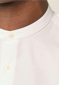 Primo piano di un collo di pelle scura che indossa una camicia bianca con colletto alla cinese e due bottoni bianchi visibili.