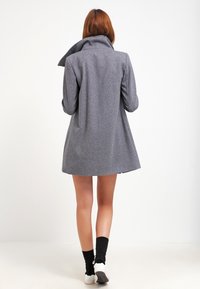 Cappotto grigio in misto lana con colletto alto e silhouette strutturata, dotato di maniche lunghe e forma a trapezio. Chiuso davanti.