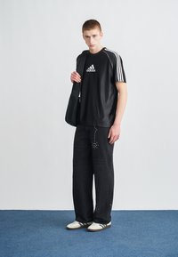 Juoda sportinė marškinėlių su balta Adidas logo ir trimis juostelėmis, derinama su laisvomis juodomis džinsais su metaliniais akcentais ir dėvėto stiliaus detalėmis.