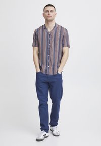 Camicia a maniche corte a righe con motivi verticali blu, rossi e bianchi, abbinata a pantaloni blu navy e sneakers bianche con dettagli neri.