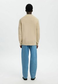Beżowy sweter dzianinowy z prążkowanymi mankietami i krawędzią, zestawiony z luźnymi, jasnoniebieskimi dżinsami. Postać stoi tyłem do widza.