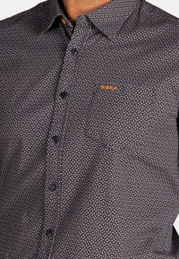 Donker button-up shirt met een micro-patroon van witte en oranje stippen. Heeft een patch pocket en subtiele branding in oranje op de borst.