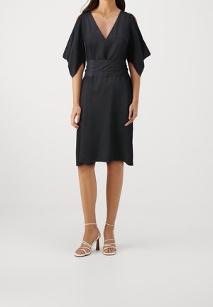 Mujer con un vestido negro hasta la rodilla, con un pronunciado escote en V, mangas cortas anchas, cintura ajustada y sandalias blancas de tacón alto con tiras.