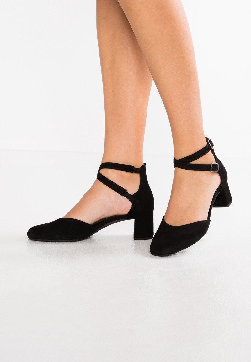 Jambes de femme portant des chaussures à talons blocs bas en daim noir avec brides et boucles à la cheville, sur un sol blanc.
