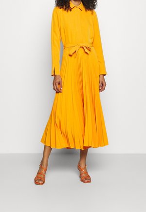 Nainen, joka pukeutuu pitkähihainen, kauluksellinen, kirkkaan oranssi pliseerattu mekko, jossa on vyötärön solmimisnauha ja oranssit remmillä varustetut korotut sandaalit, seisoo valkoista taustaa vasten.