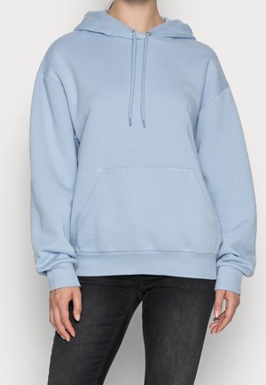 Person iført lyseblå hættetrøje med frontlomme og sorte tætsiddende jeans, stående mod en ensfarvet baggrund.
