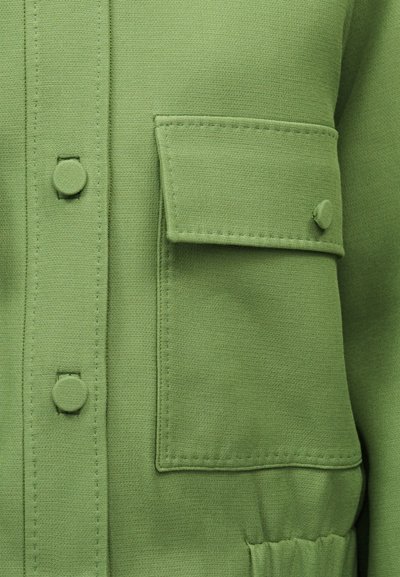 Gros plan sur une veste verte texturée montrant une poche à rabat boutonnée et des boutons recouverts de tissu le long de la patte de boutonnage avant.