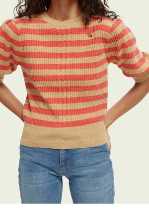 Nainen, jolla on beige ja korallinvärinen raidallinen lyhythihainen neulepusero, jossa on kierretty neulekuvio, yhdistettynä sinisiin farkkuihin, kädet lanteilla.