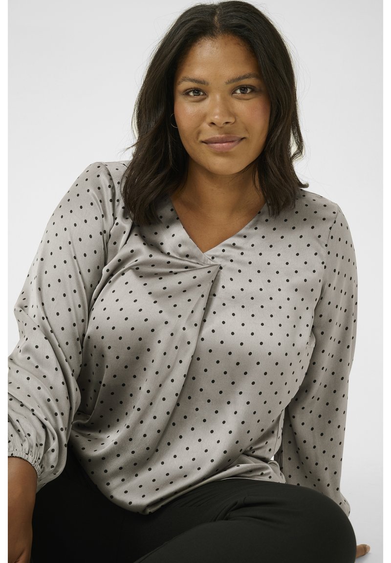 Blouse grise soyeuse à pois noirs, décolleté en V, détail froncé à l'avant et manches élastiquées. Associée à un pantalon noir.