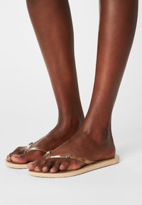 Tongs à brides transparentes avec une semelle plate en tan. Les brides sont dotées de détails délicats en matériel pour un ajout de style.