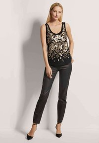 Zwarte mouwloze top met gouden bloemenborduursel, gecombineerd met zwarte slim-fit broek en pumps met spitse neuzen en enkelbandjes.