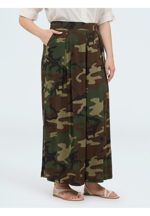 Persoon die een lange geplisseerde camouflage rok, een wit shirt met korte mouwen en beige sandalen draagt, staat met een hand in de zak.