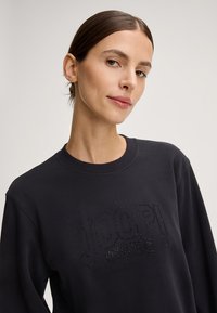 Sweatshirt grigio scuro con scollatura a coste e maniche lunghe, con un logo testurizzato "JOOP! JEANS" in ornamenti scuri sul davanti.