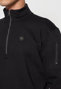 Zwarte pullover met rits en hoge kraag, voorzien van een hexagonaal logo en een ritssluiting op de mouw. Gladde textuur, moderne pasvorm.