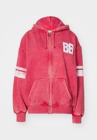 Sudadera roja con cremallera hecha de tela texturizada. Presenta un estampado blanco de "86", franjas blancas en las mangas y un bolsillo frontal.