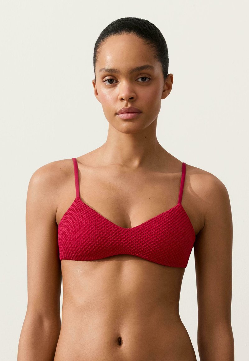 Femme aux cheveux foncés tirés en arrière, portant un haut de bikini rouge texturé avec des bretelles fines, debout devant un fond clair uni.