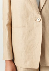 Blazer en lin beige avec une fermeture à un bouton, un col à revers et une poche latérale. Le tissu présente une finition lisse et texturée.