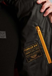 Main tenant une veste noire ouverte montrant une fermeture éclair jaune et une patch noire portant l'inscription "GOBBLER BW ISSUE 508/00 DRAWING NO. PB13 PME LEGEND TESTED AT HIGH ALTITUDE."