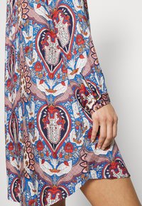 Lähikuva kädestä, joka lepää sinisen, punaisen ja valkoisen kukkakuvioisen mekon päällä. Mekossa on pitkät hihat ja napillinen kalvosin.