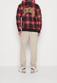 Felpa a quadri rossi e neri con la scritta "JORDAN" sulla schiena, abbinata a pantaloni della tuta beige e sneakers bianche con accenti rossi.