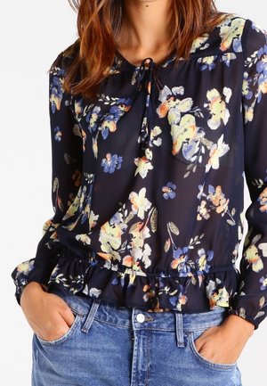 Marineblouse met geel, wit en paars bloemmotief, lange mouwen, gerimpelde zoom, gestrikte halslijn, gedragen en ingestopt in een blauwe spijkerbroek.