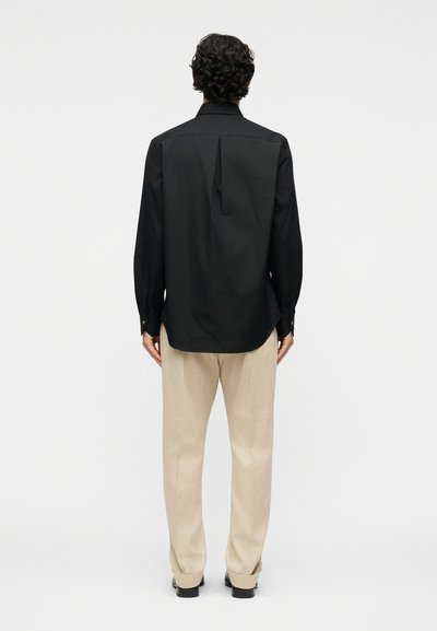 Άνδρας με σγουρά μαλλιά που κοιτάει προς τα πίσω, φοράει μαύρο μακρυμάνικο πουκάμισο και μπεζ παντελόνι μπροστά σε απλό φόντο.