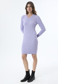 V-Ausschnitt, langärmliges Kleid aus Kabelstrick in hellem Lavendel. Kombiniert mit schwarzen Stiefeletten, die eine figurbetonte Silhouette und gerippte Textur zur Geltung bringen.