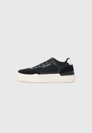 Zwarte sneaker van mesh en leer, met een dikke witte zool en contrasterende accenten. Veters en logodetail aan de zijkant.