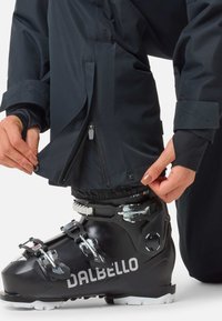 Czarne buty narciarskie z logo Dalbello, z metalowymi klamrami i teksturowaną powierzchnią. Dostosowywana jest wodoodporna kurtka zapinana na zamek.