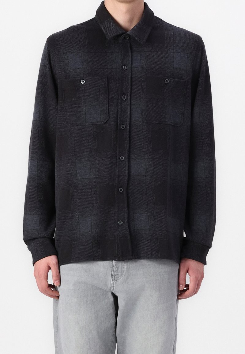 Homme portant une chemise noire à manches longues boutonnée avec deux poches poitrine et un pantalon gris clair, debout devant un fond uni.