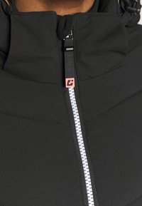 Gros plan sur une fermeture éclair de veste noire avec une languette de tirage noire et un petit logo carré rouge sur le cou et la poitrine supérieure d'une personne.