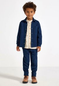 Laivastonsininen paita takki, jossa kaksi rintataskua, päällä kerma nappipaita, jossa on pieniä ruskeita kuvioita. Yhdenmukaiset laivastonsiniset housut ja ruskeat saappaat.