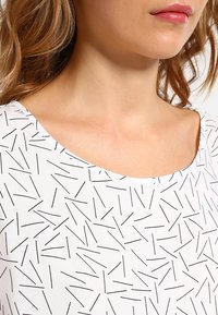 Chemise blanche à encolure ronde large, ornée d'un motif géométrique noir. Tissu léger, texture lisse, design décontracté.