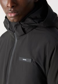 Veste imperméable noire avec un col haut, dotée d'une fermeture éclair sur le devant, d'une capuche avec cordon de serrage, et d'un petit logo Calvin Klein blanc sur la poitrine.