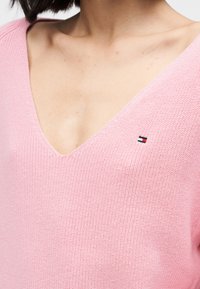 Ανοιχτό ροζ πουλόβερ με V λαιμόκοψη από υφασμάτινο πλεκτό με υφή, που διαθέτει ένα μικρό λογότυπο στην αριστερή πλευρά του στήθους.