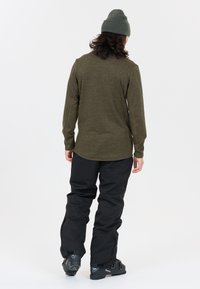 Oliven grøn langærmet skjorte med en struktureret overflade, parret med sorte snebukser. Hovedet delvist dækket af en grå strikket hue.