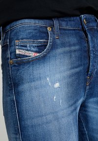 Tmavě modré džíny z denimu s bledým umytím, s poškozenou oblastí na stehně, malou přední kapsou a značkovým štítkem poblíž kapsy.