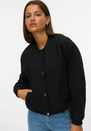Femme portant une veste bomber noire à boutons avec des poignets et un col côtelés, une main dans la poche, associée à un jean bleu clair.