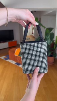 Hender som holder en liten svart veske med et sølv rhinestones rutenettmønster og svarte håndtak i en stue.