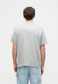 Άνδρας με κοντά καστανά μαλλιά που φοράει απλό γκρι κοντομάνικο μπλουζάκι και ανοιχτό μπλε τζιν, στέκεται με την πλάτη προς τα εμπρός σε λευκό φόντο.