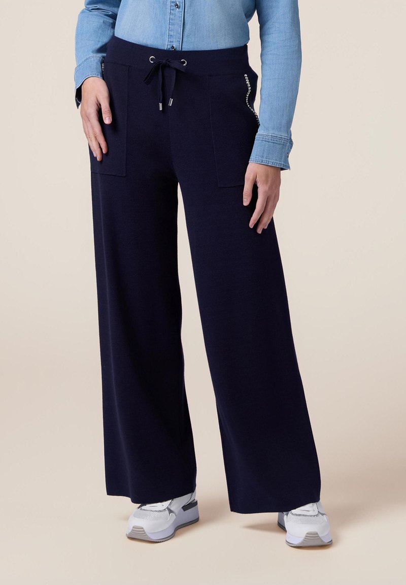 Pantaloni a gamba larga blu navy con due tasche frontali, cintura con cordino e dettagli argentati sottili, abbinati a scarpe da ginnastica bianche.