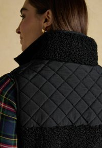 Czarna pikowana kamizelka z teksturowanym kołnierzem z polaru. Projekt zawiera kratkowany wzór na spodniej koszuli, widocznej z boku.