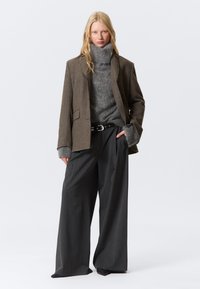 Overmaatse bruine blazer met een gestructureerde afwerking over een grijze coltrui en losse grijze broek. Zwarte riem met zilveren sluiting als accent.