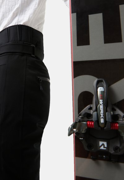 Czarne spodnie narciarskie z fakturowanym pasem i zamykaną na zamek kieszenią, zestawione z deską snowboardową o ciemnym wzorze i czerwonymi akcentami.