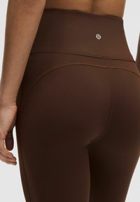 Hochtaillierte braune Leggings aus dehnbarem Material, mit nahtlosem Design und einem kleinen Logo in der Nähe des Bundes. Glatte Textur.
