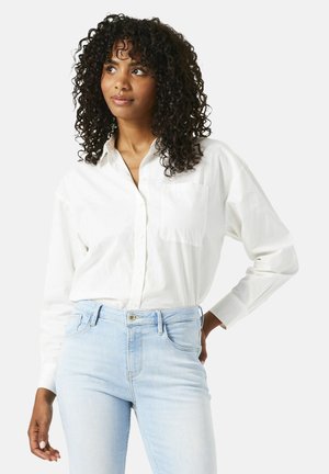Chemise blanche à manches longues avec un unique poche poitrine, associée à un jean ajusté bleu clair. Texture douce et coupe décontractée.
