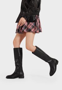 Stivali neri fino al ginocchio con una texture in pelle liscia, punta rotonda, suola piatta e dettagli con zip laterale, su uno sfondo semplice.