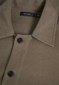 Khaki színű, gombbal záródó ing, textúrált anyagból, széles gallérral és elöl két fekete gombbal. A márka címkéje "Matinique".